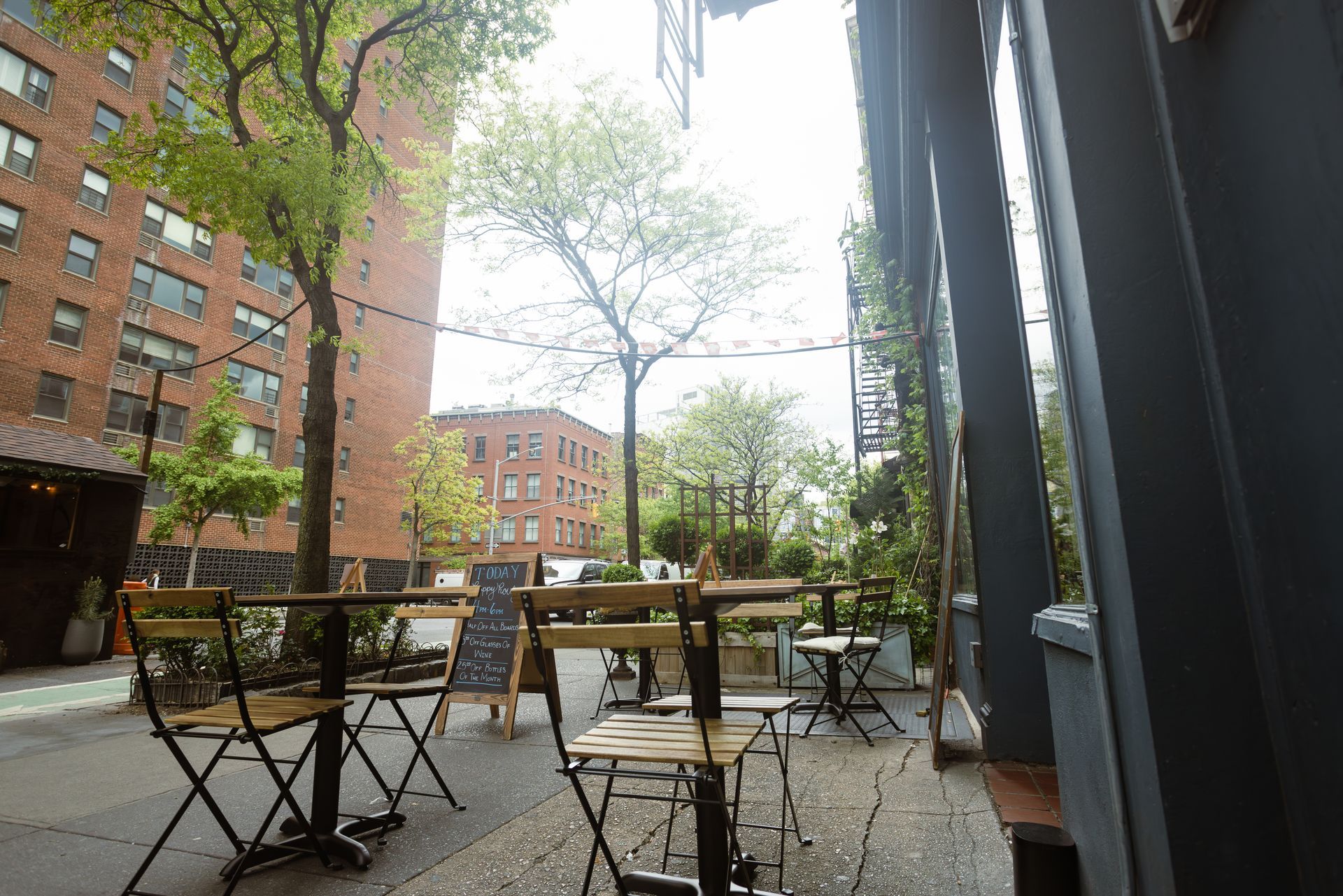 Outdoor café patio with metal tables and chairs beside brick apartments on a tree-lined street