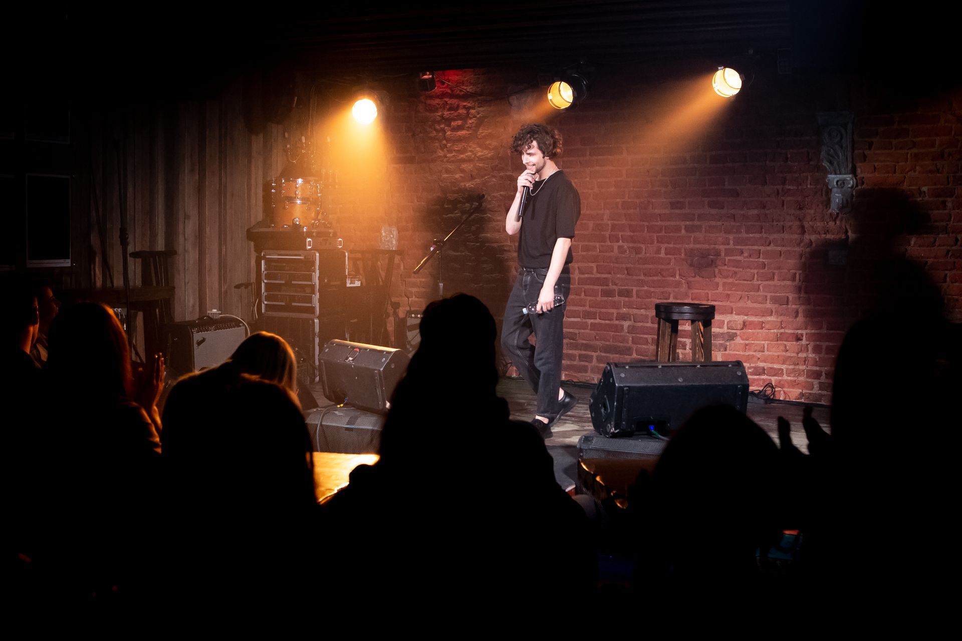 Comedian performing on a small stage under warm spotlights, with audience silhouettes in front.