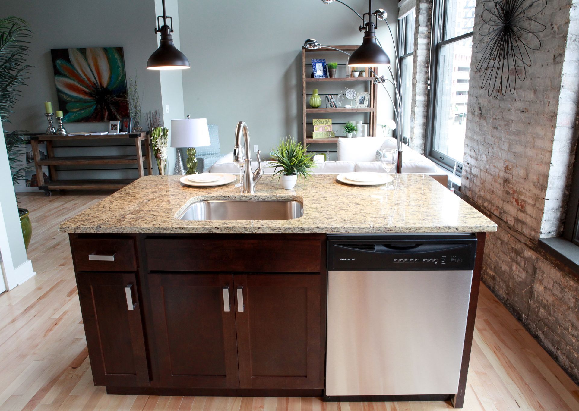 Modern kitchen island with granite countertop, sink, dark cabinets, and stainless dishwasher by a brick wall.