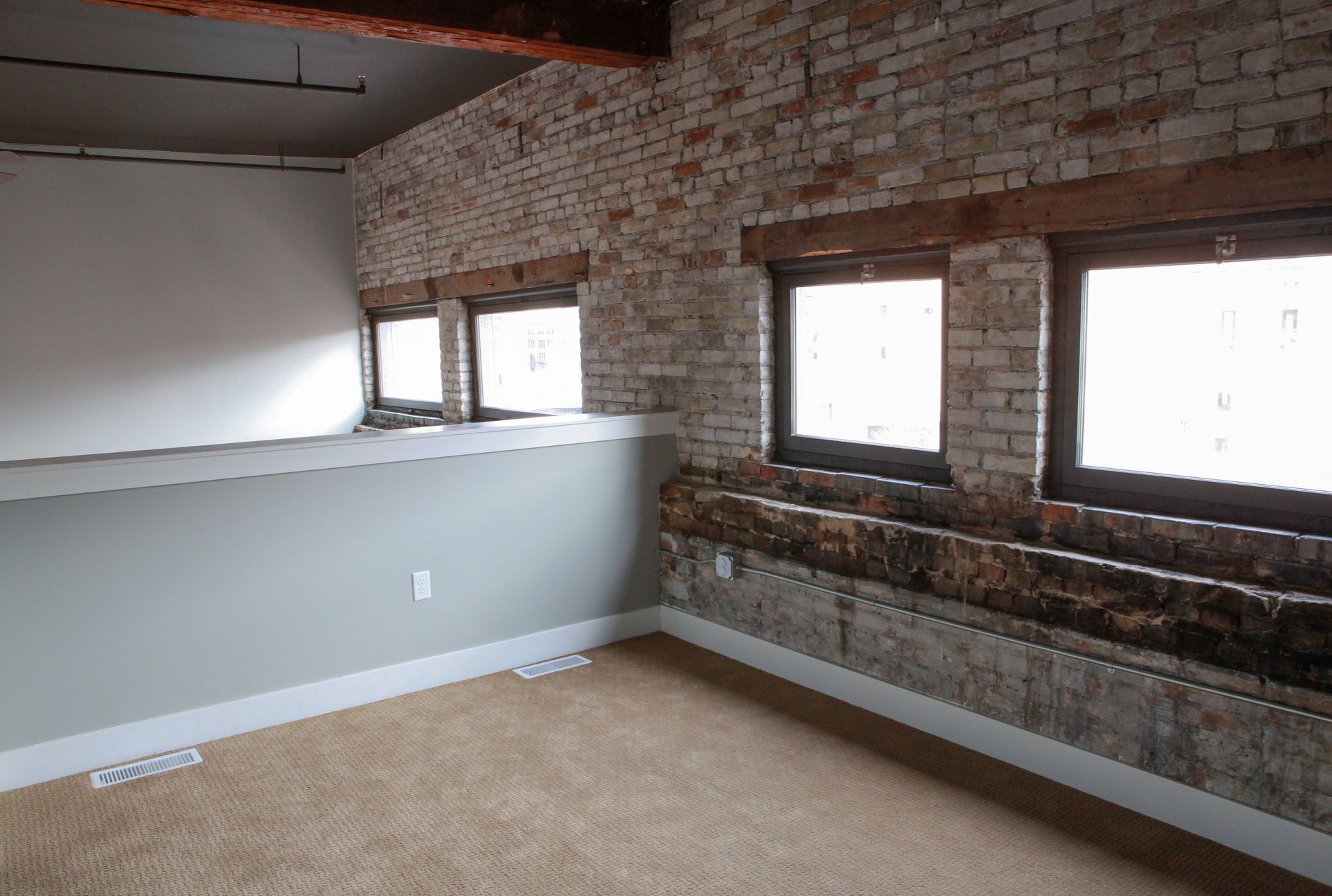 Loft-style room with exposed brick walls, three windows, gray half-wall, and beige carpet.