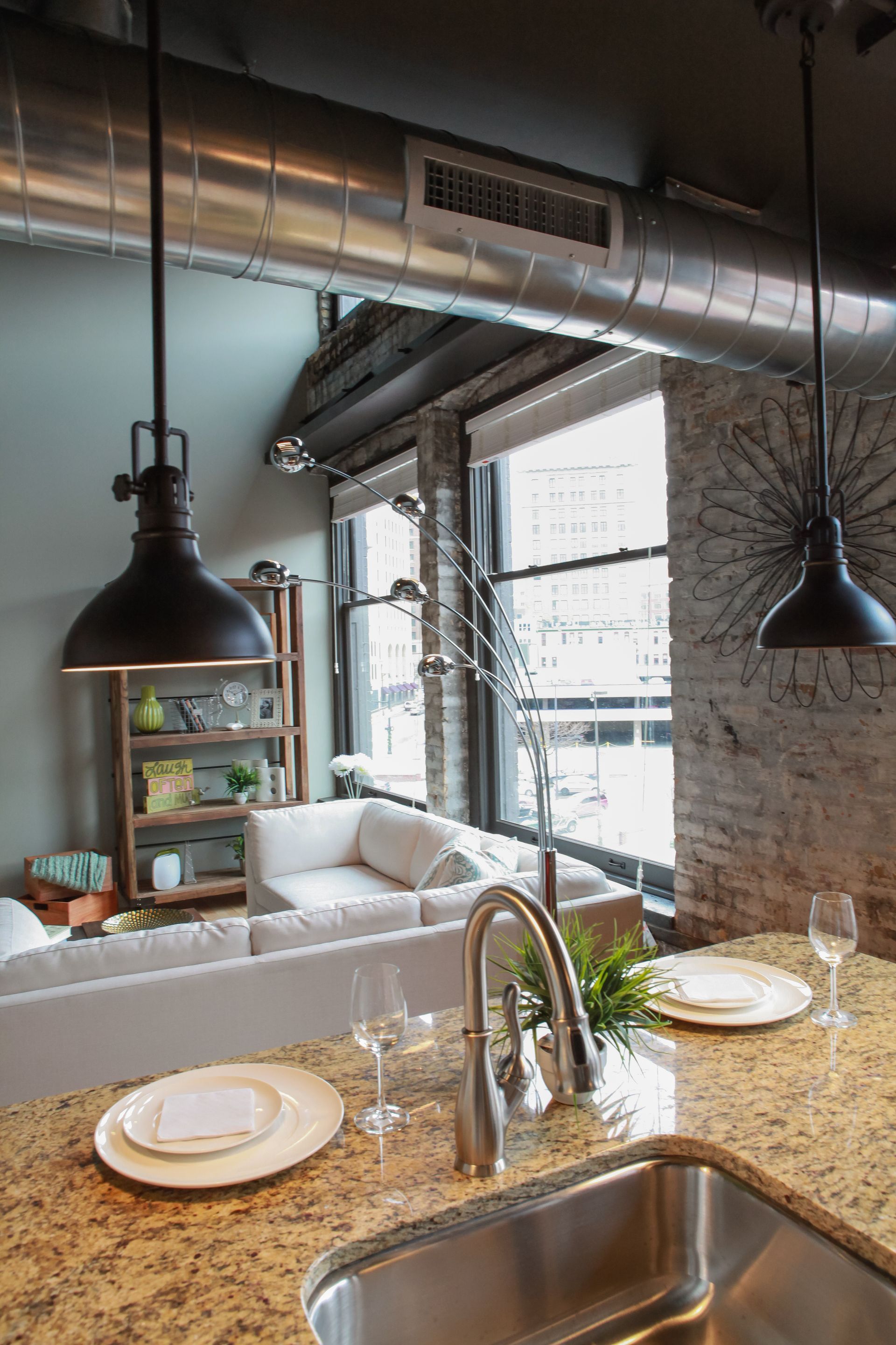 Modern kitchen with granite island, sink, pendant lights, exposed brick, and large windows overlooking a living area