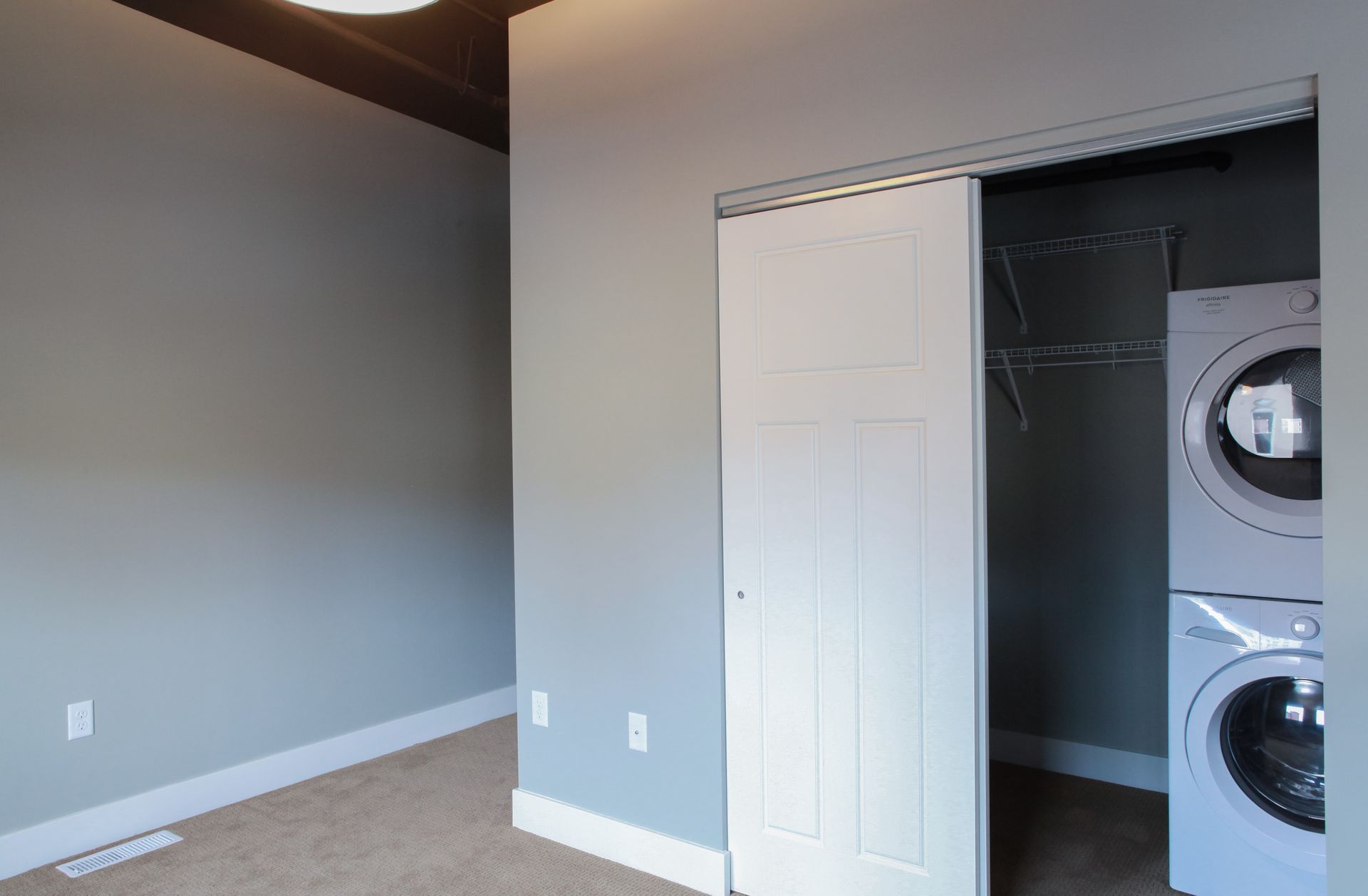Empty bedroom with gray walls, beige carpet, and an open closet with stacked washer and dryer.