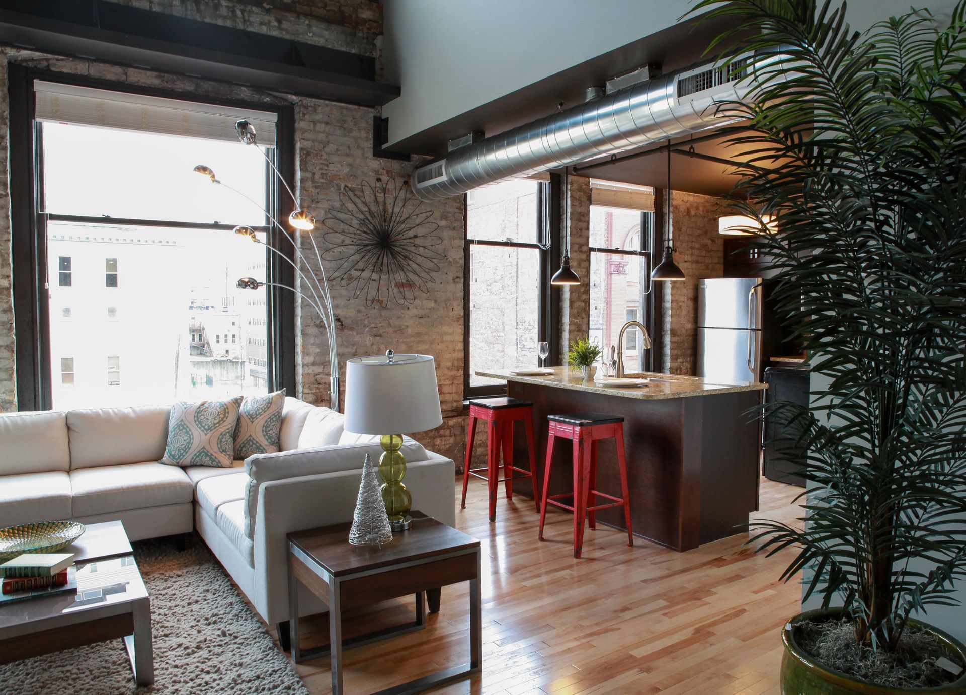 Bright loft living room with white sectional, wood table, red stools, exposed brick, and large windows