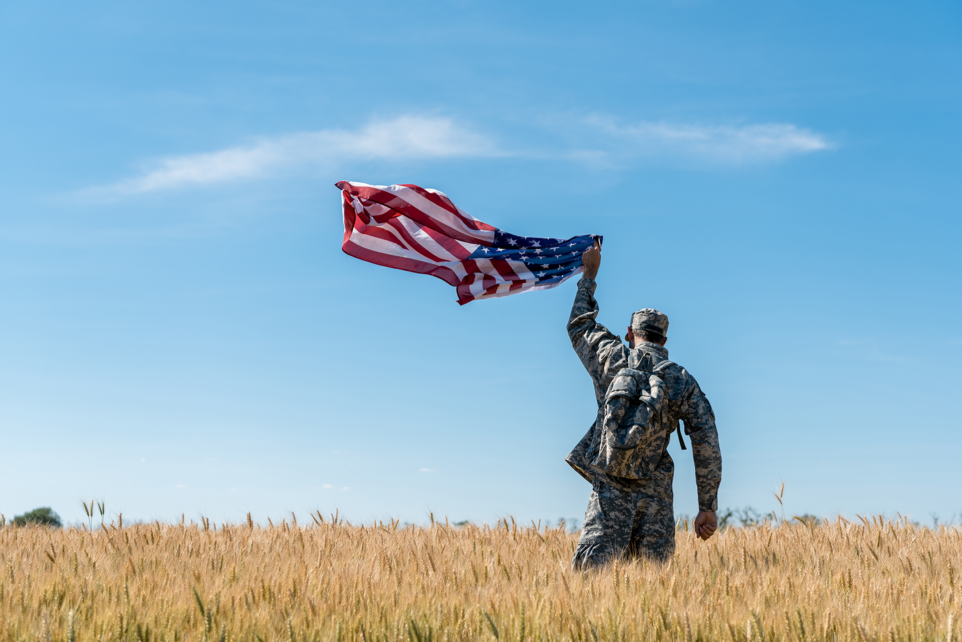 Soldier in camouflage holding up an American flag in a wheat field under a blue sky.
