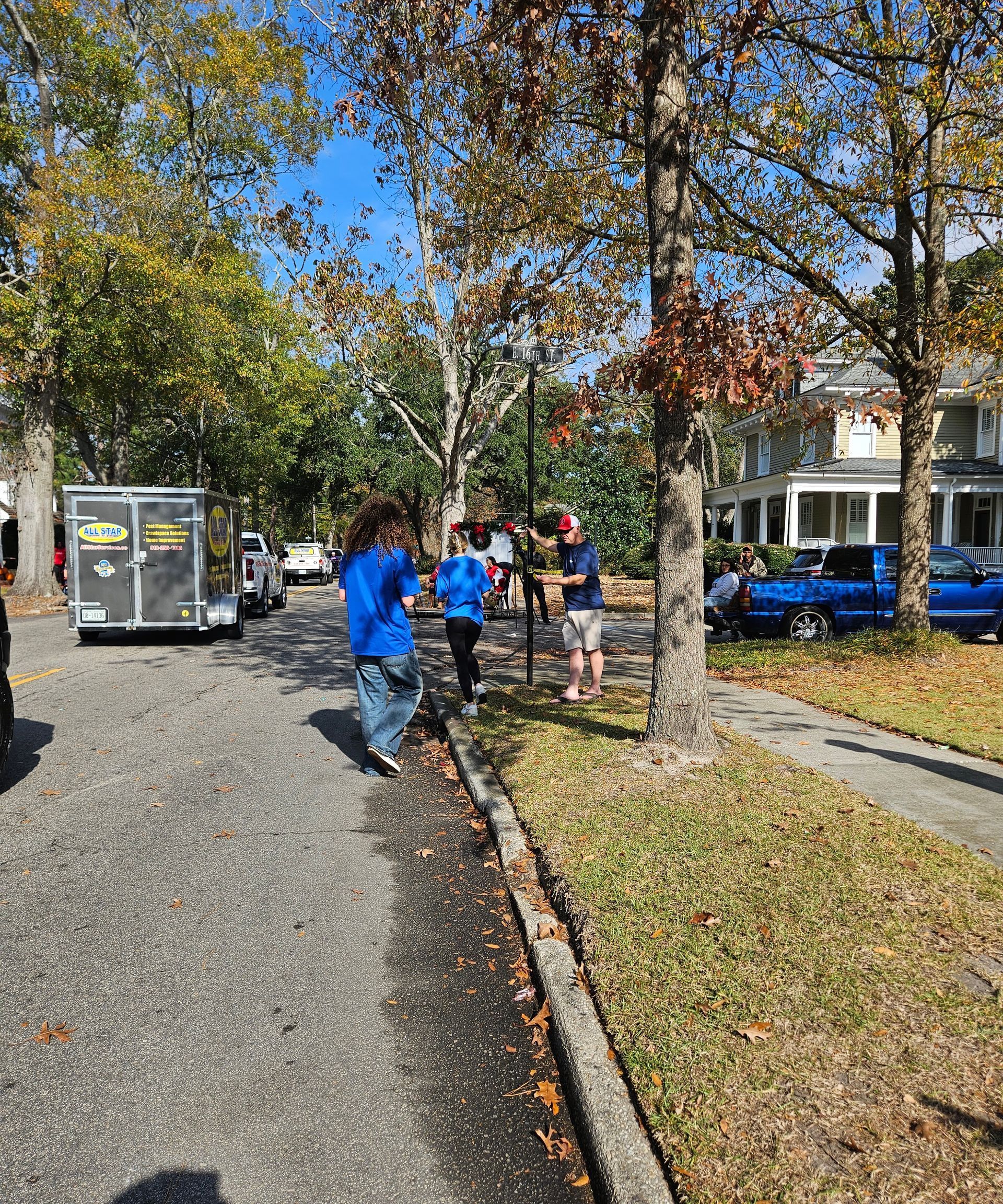 Three people stand near a trailer and a blue truck on a tree-lined residential street on a sunny day.