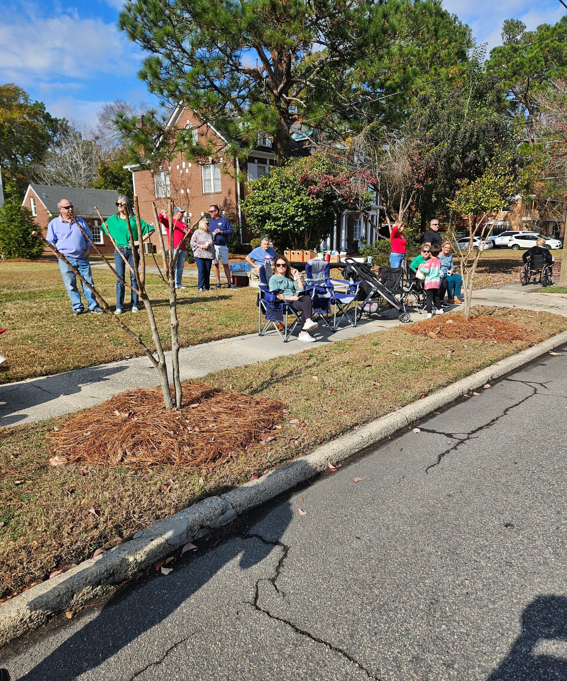 A group of people standing and sitting in lawn chairs on a residential lawn on a sunny day.
