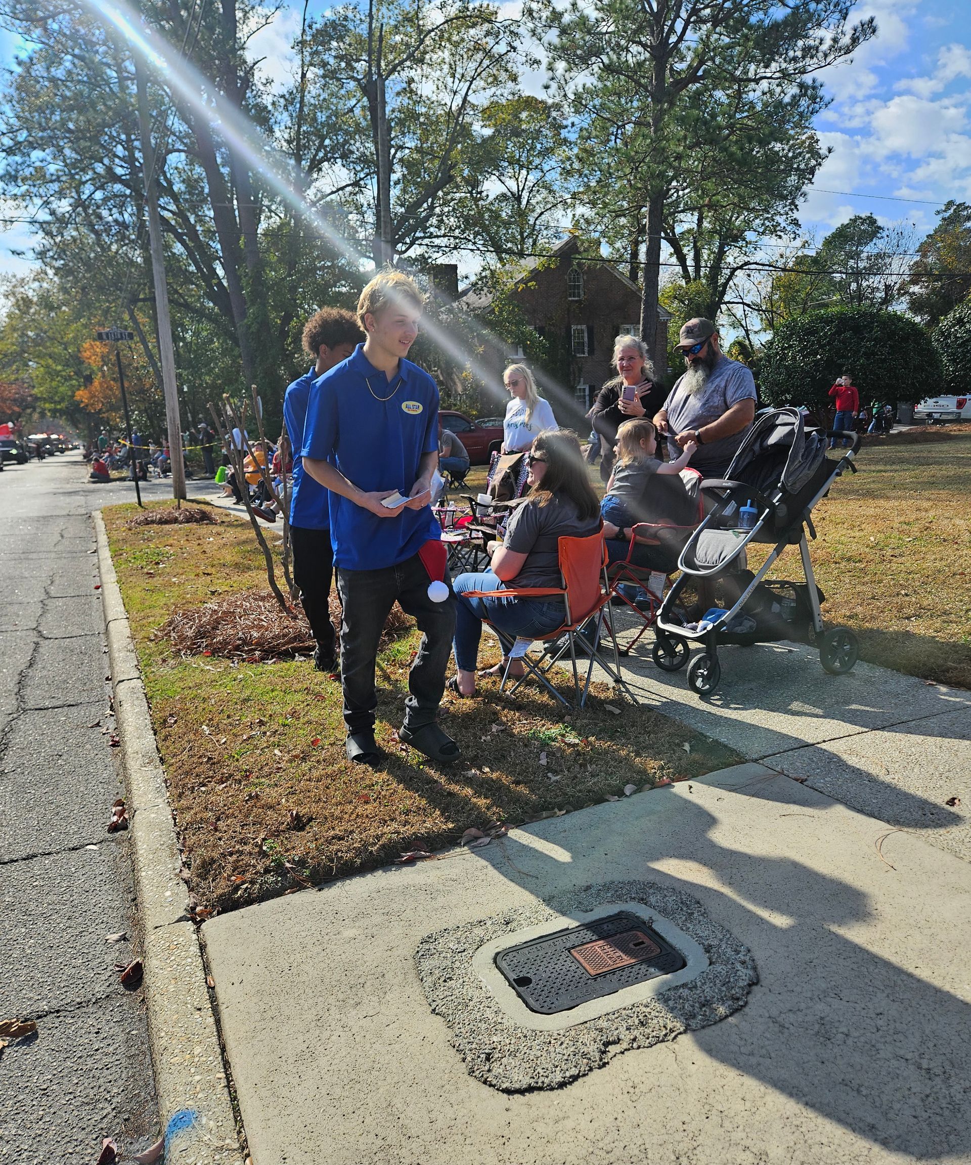 People wearing blue shirts stand or sit on a grassy roadside, some with folding chairs, on a sunny day.