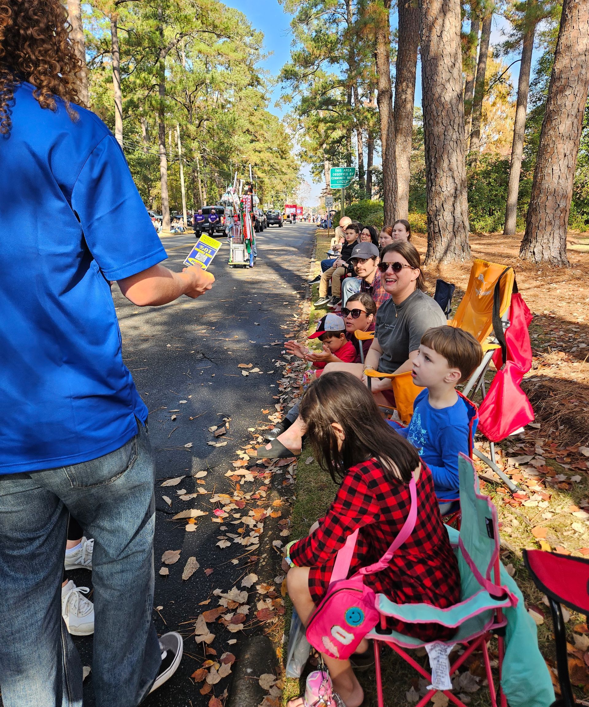People sit on camping chairs along a road, watching a parade while a person in a blue shirt stands in the foreground.