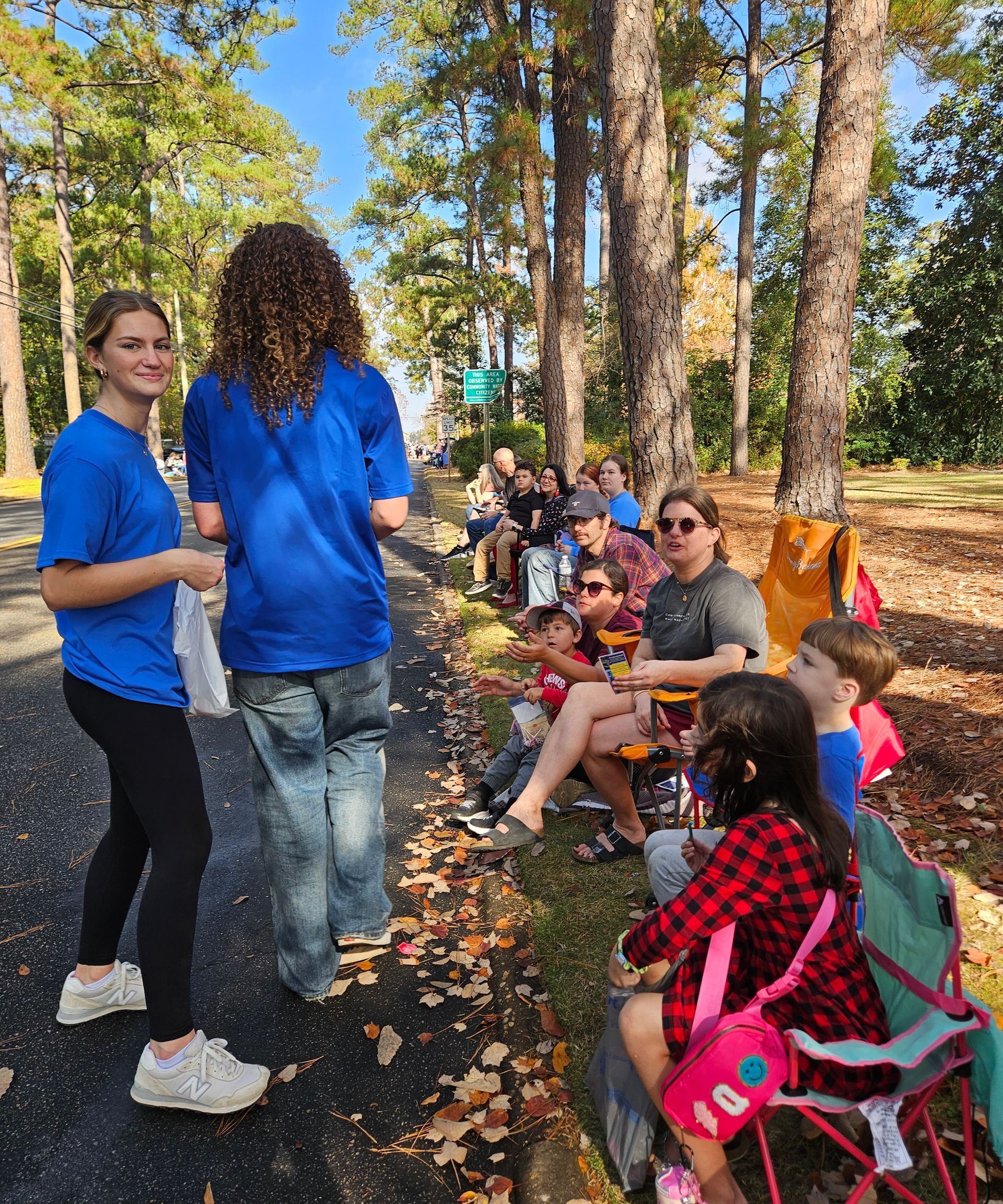 Two people in blue shirts stand beside a roadside gathering of people sitting in folding chairs under trees.