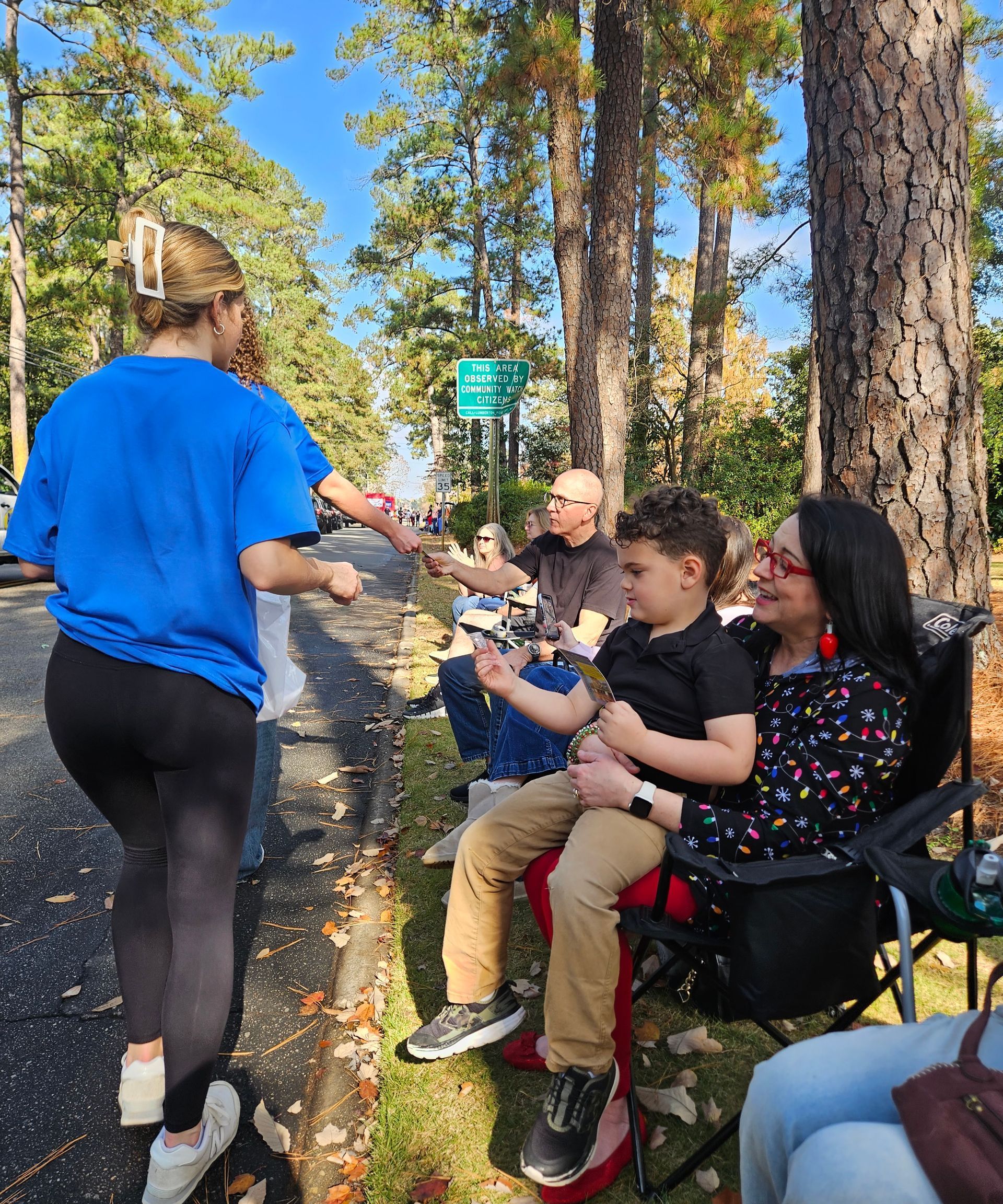 A person in a blue shirt handing out items to spectators sitting on the side of a road lined with trees.