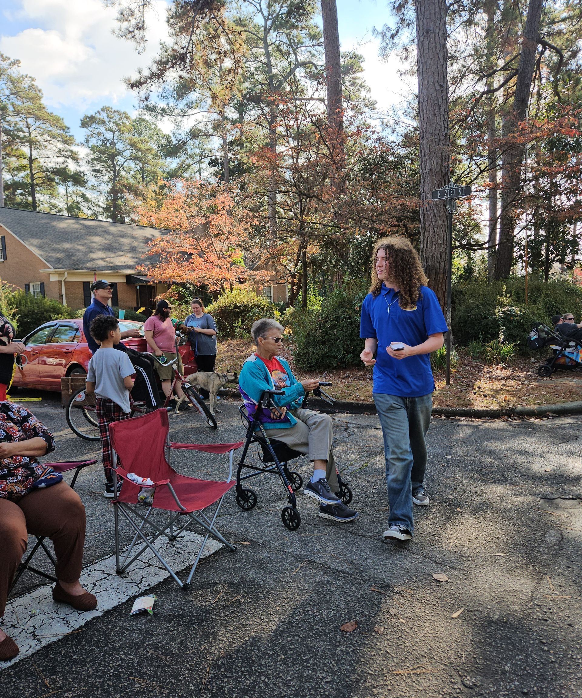 People gather outdoors in a wooded area near a home, with a person standing by someone seated in a walker.