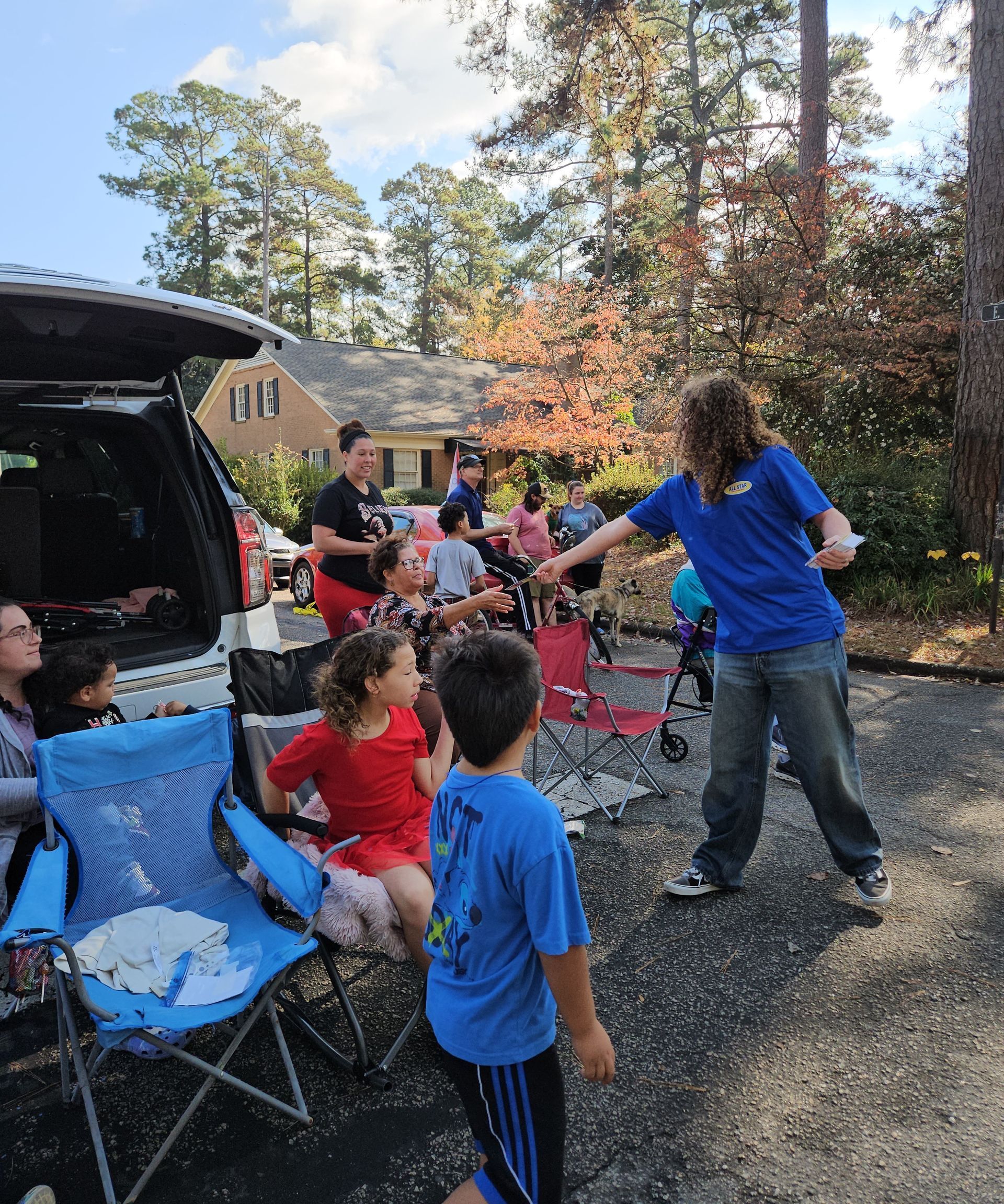 A group of people gathers near a vehicle in a wooded outdoor area, with a person in a blue shirt reaching out.