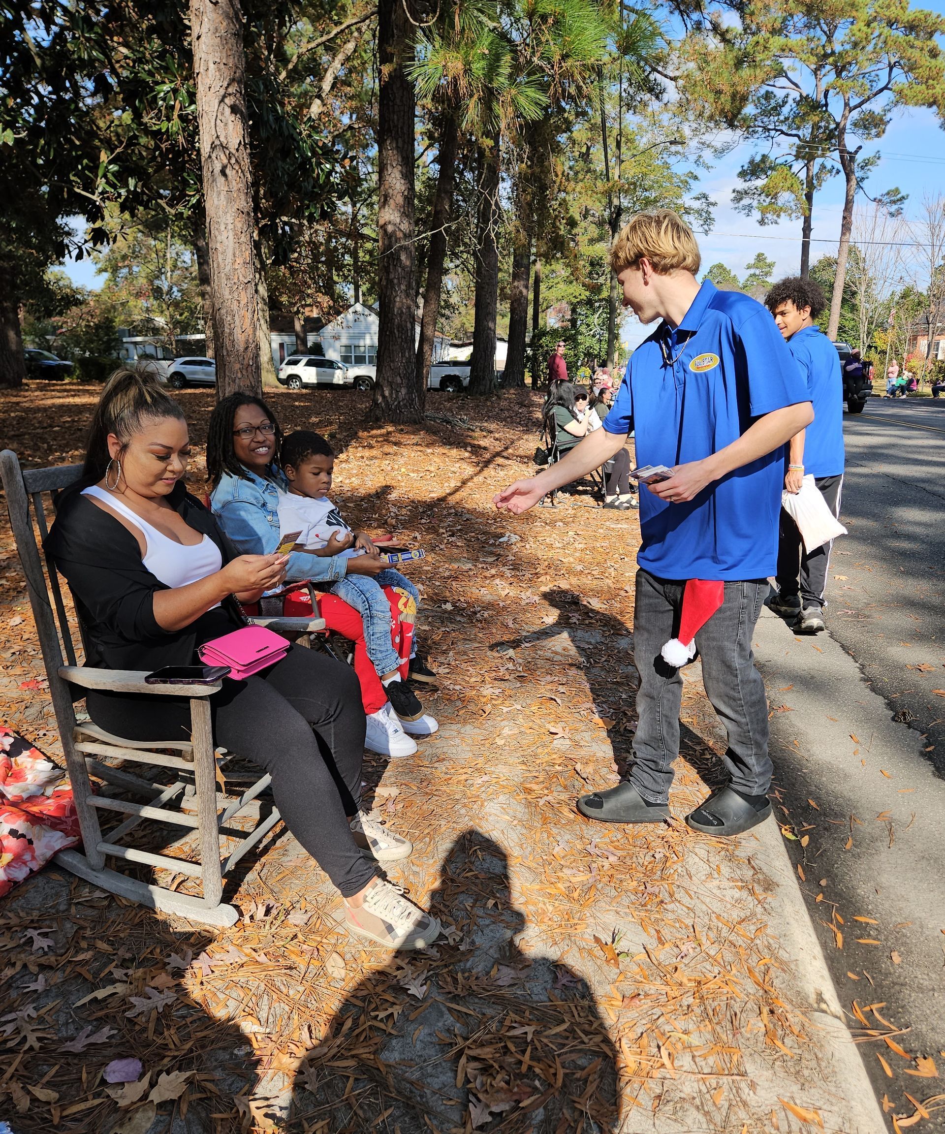 A person in a blue shirt hands a gift to a small child sitting with two adults in a park with fallen leaves.