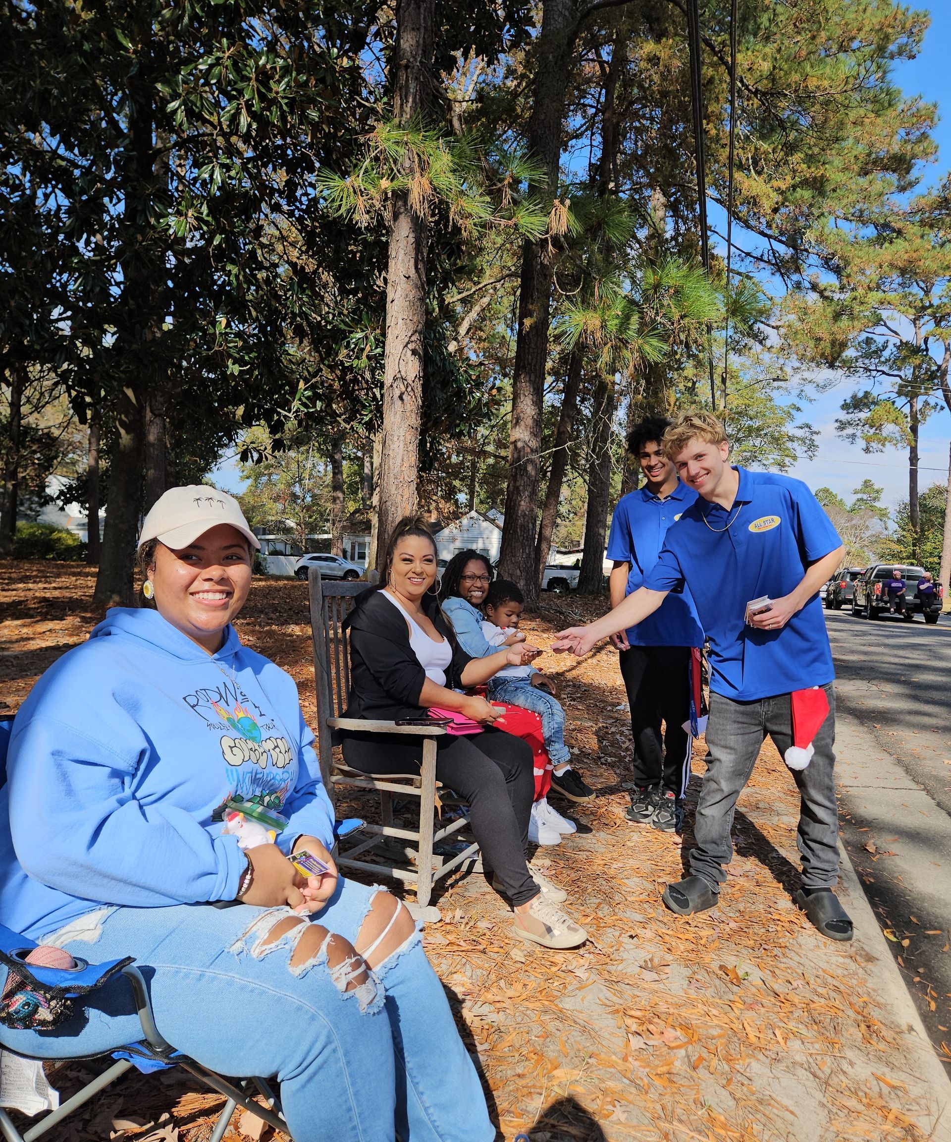 A person in a blue hoodie sits in a chair outdoors as a person in a blue shirt hands an object to a seated woman nearby.