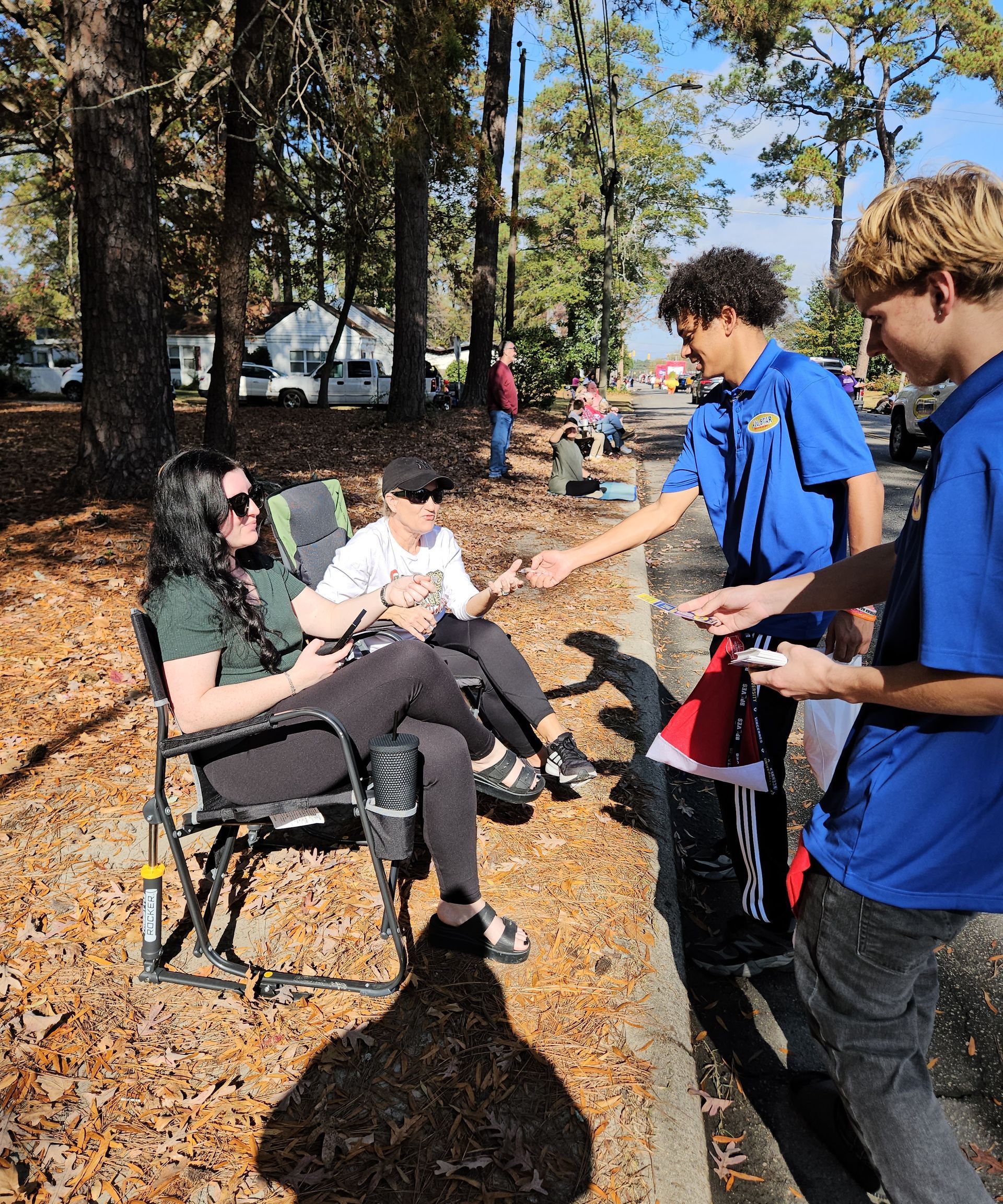 Two people in blue shirts hand items to two people seated in folding chairs along a tree-lined street covered in leaves.