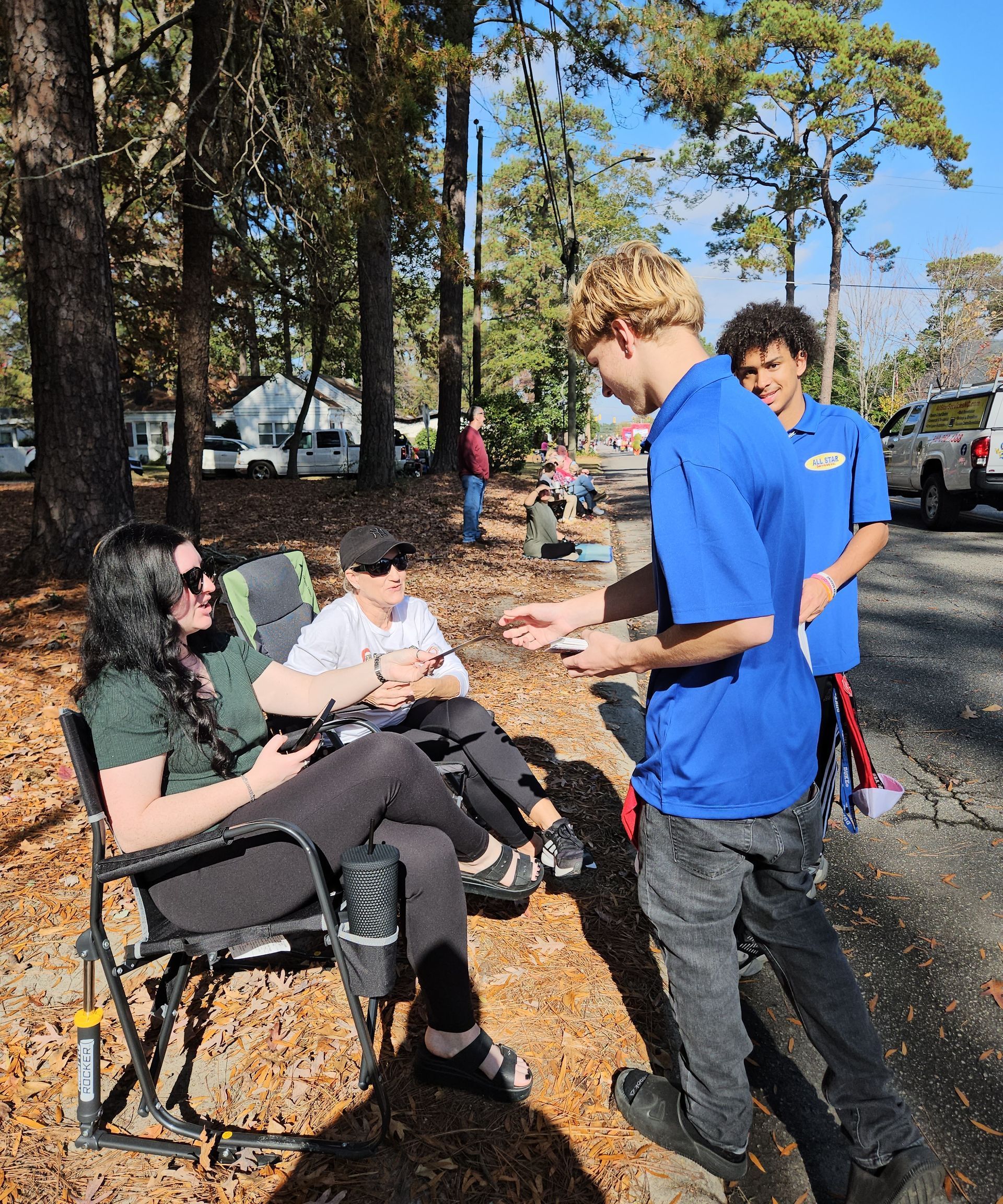 Two people in blue shirts interact with two others seated in folding chairs along a tree-lined, sunlit roadside.