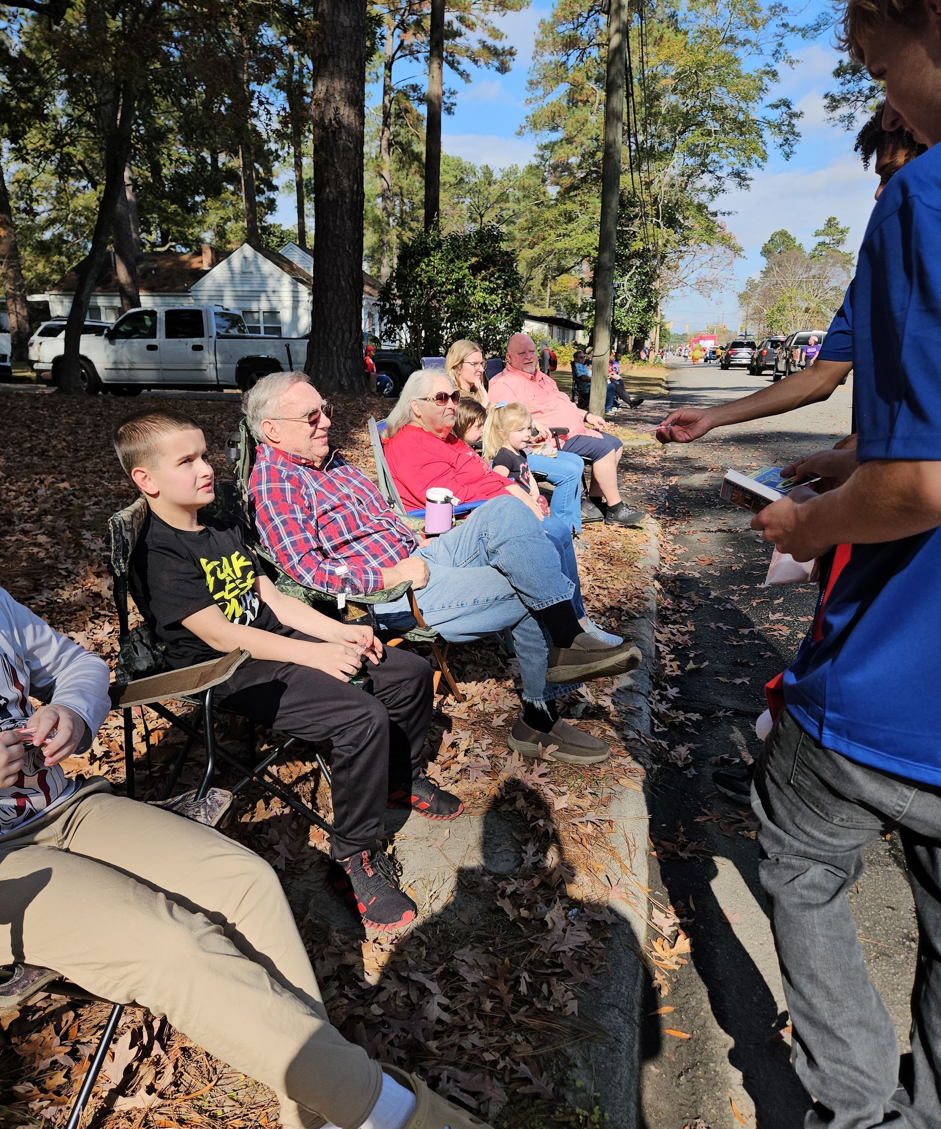 A group of people sitting in lawn chairs on a leaf-covered roadside, with a person standing and handing something out.