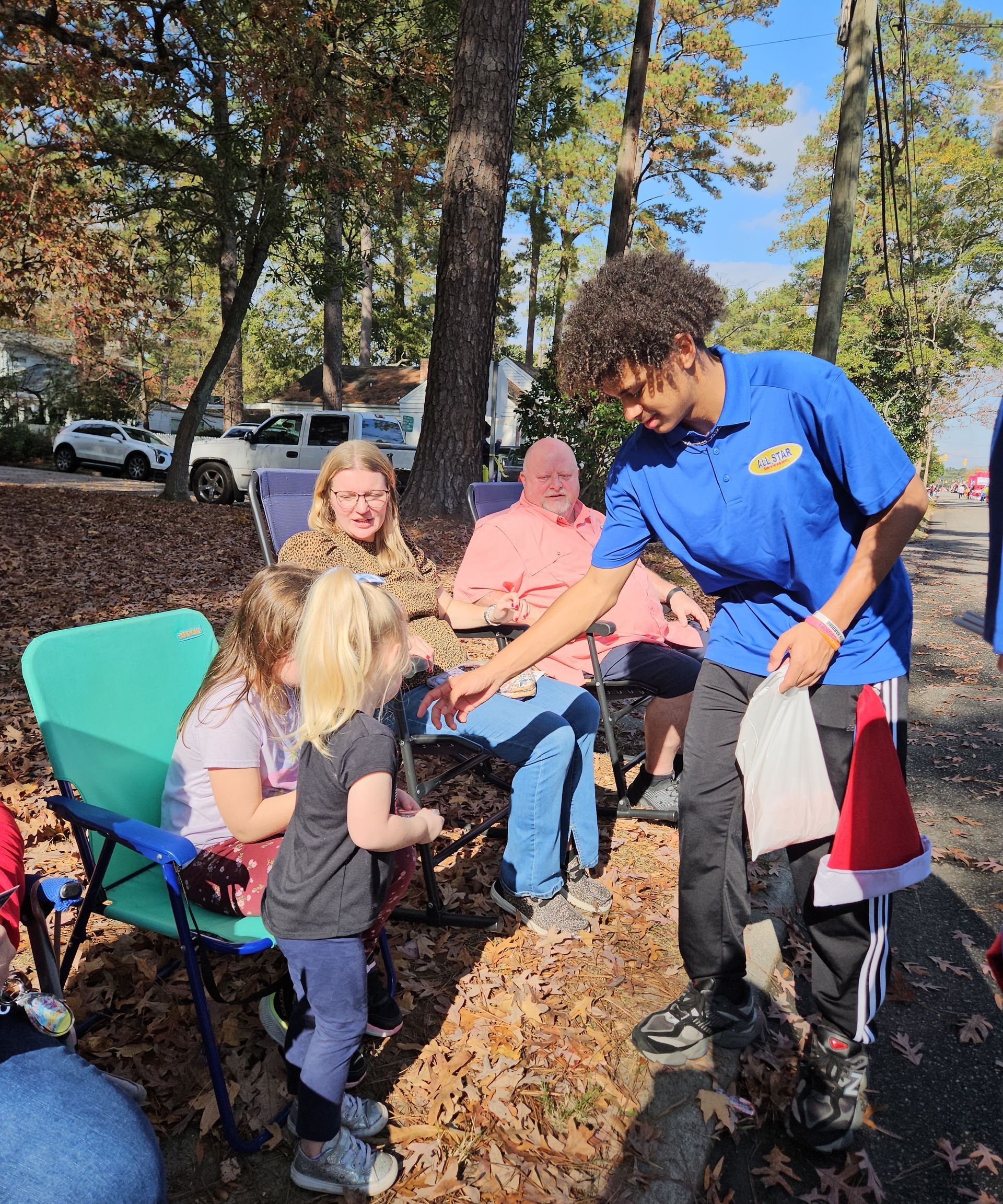 A person in a blue shirt hands a gift to a young child at an outdoor gathering among trees in an area covered with leaves.