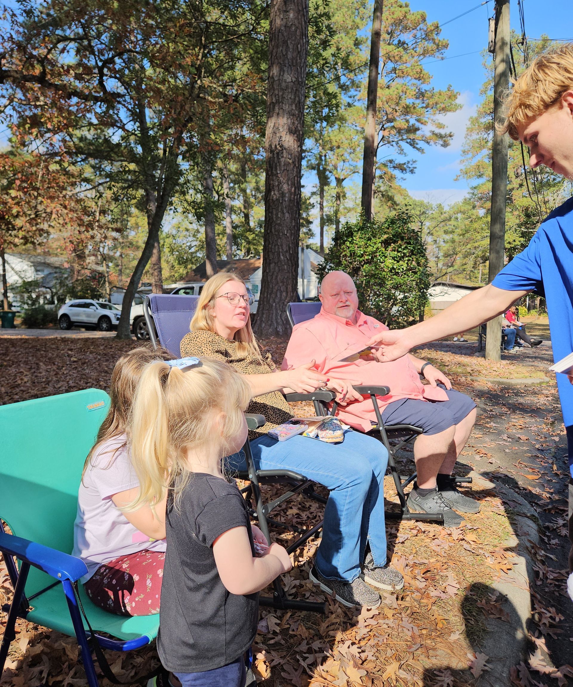 A person hands a small object to another person while seated outdoors, with children and others nearby in a wooded area.