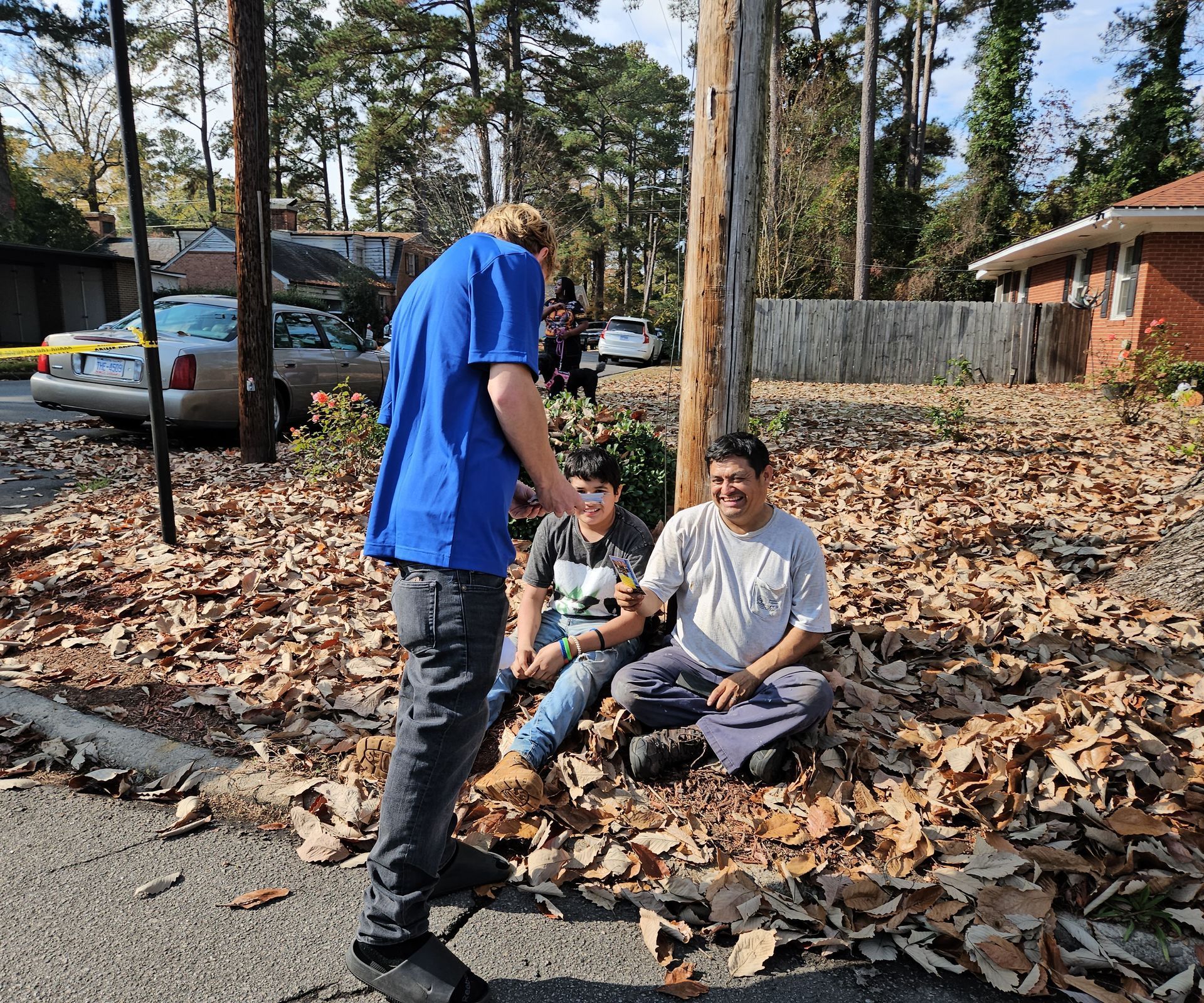 Three people interacting outdoors amidst fallen leaves near a residential street, with a car and utility pole in the back.