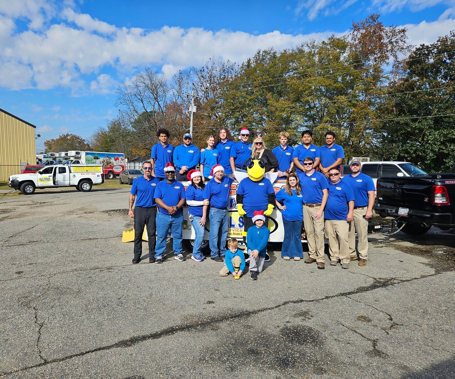 A group of people wearing blue shirts pose outdoors in a sunny lot with a mascot, some wearing festive Santa hats.
