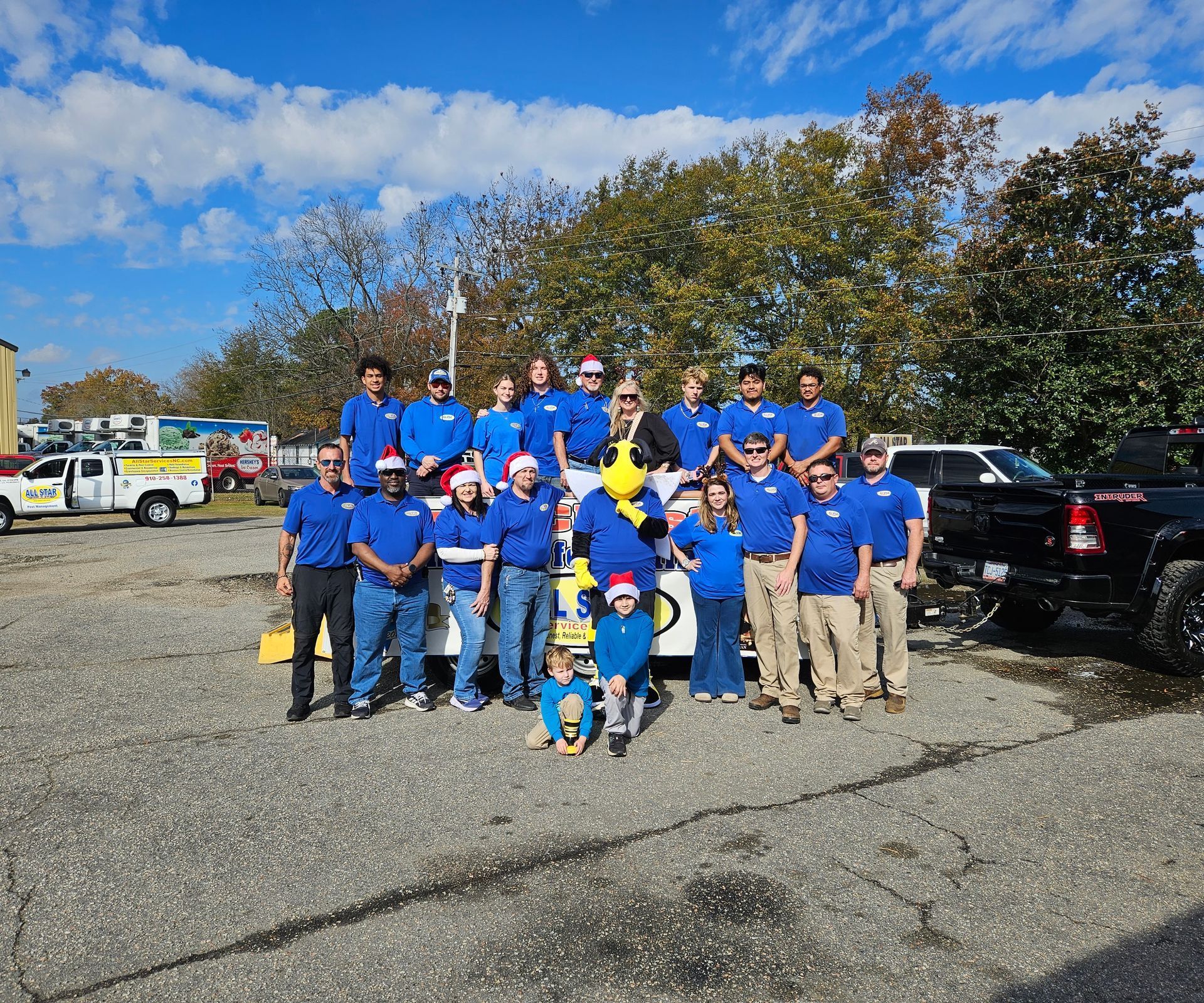 A group of people, including someone in a mascot costume, pose together outdoors on a sunny, paved lot.