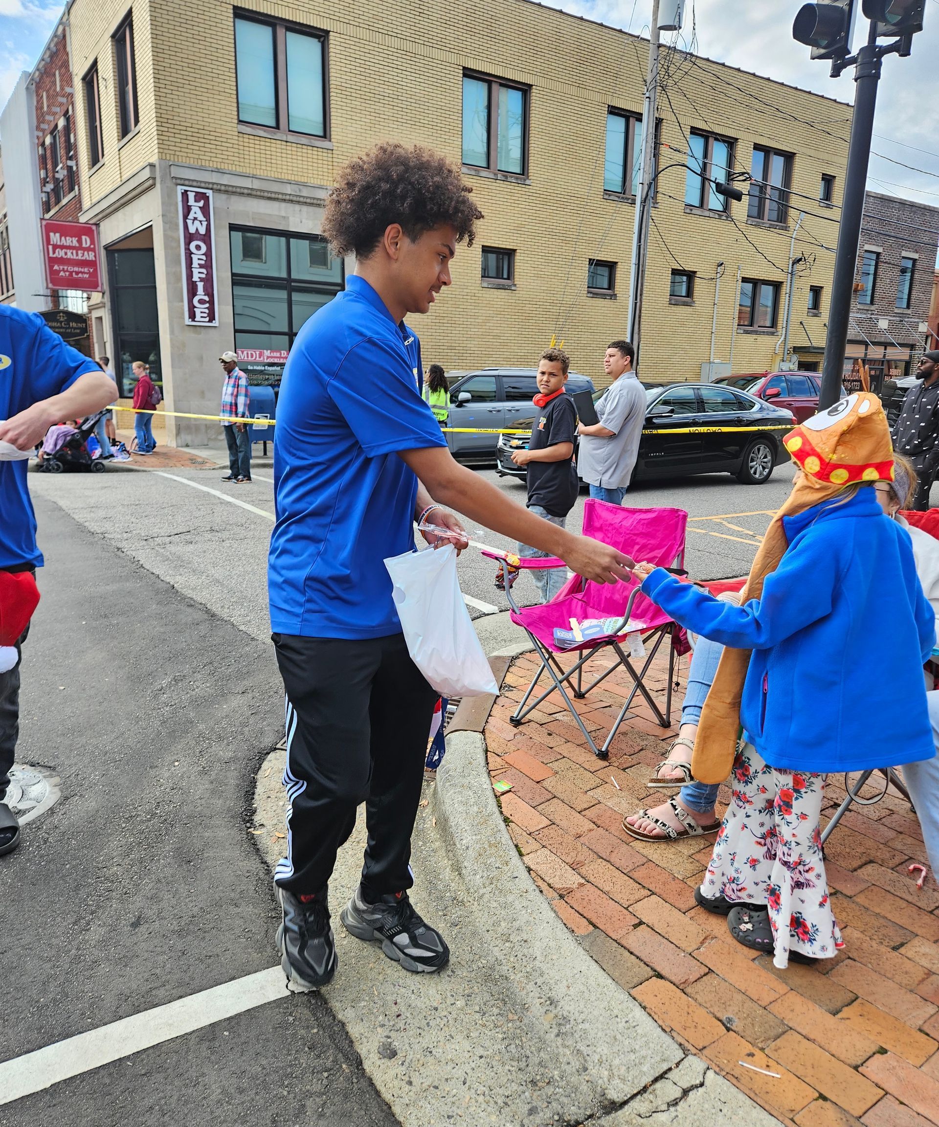 A young person in a blue shirt hands a treat to someone wearing a colorful costume on a brick sidewalk during a parade.