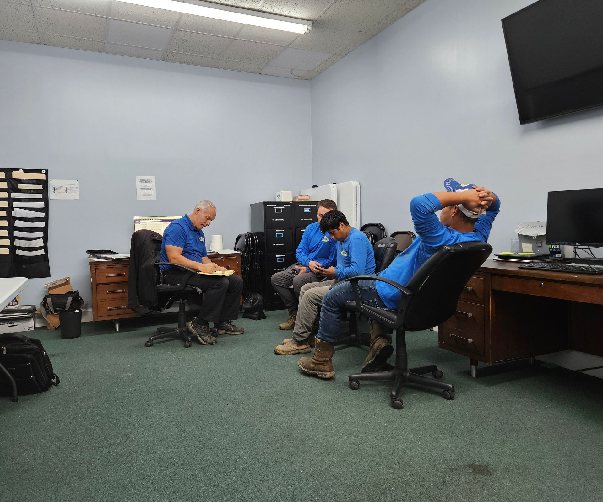 Three people in blue shirts are sitting and relaxing in a room with office desks, chairs, and a wall-mounted television.