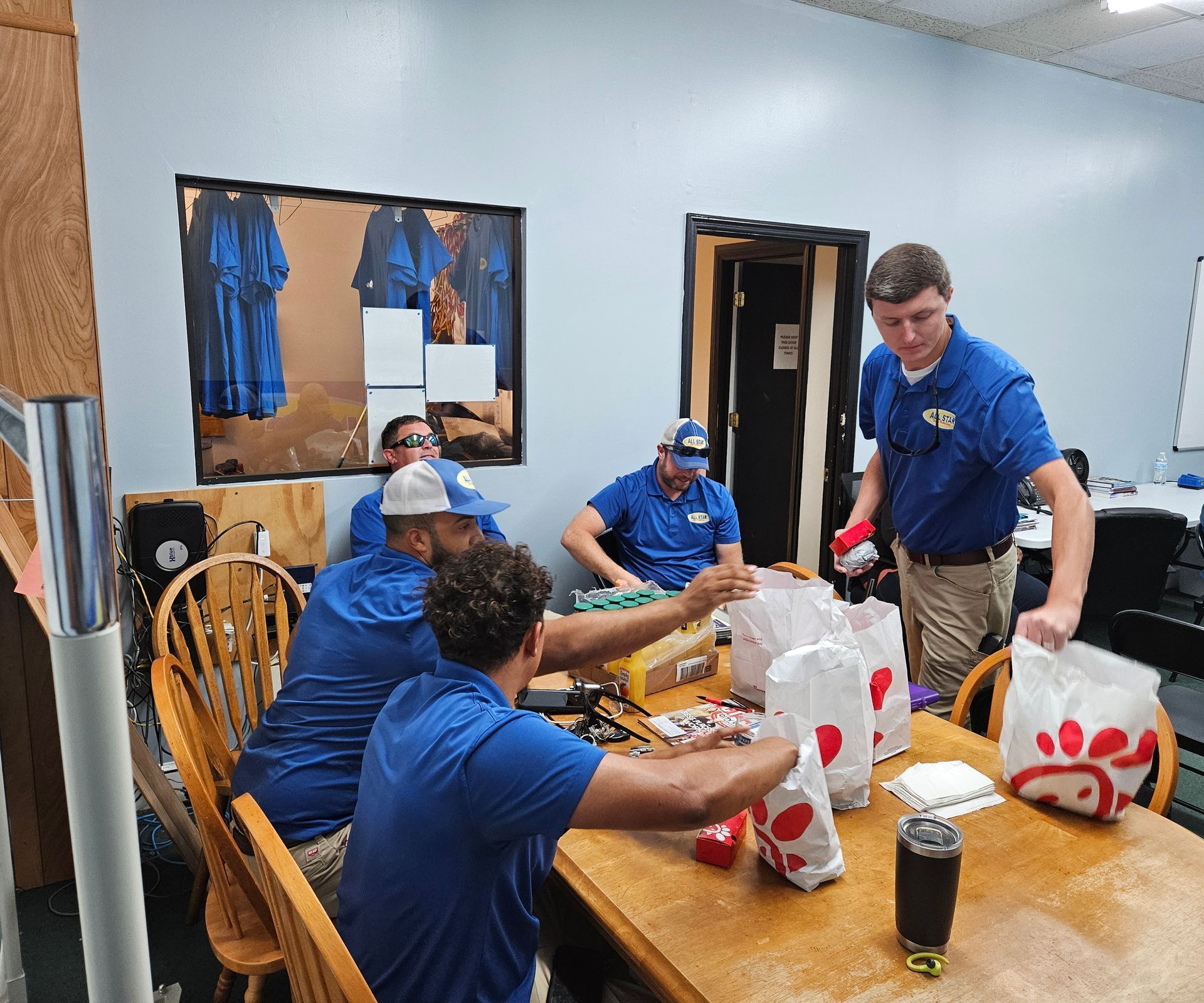 A group of people wearing blue uniforms pack Chick-fil-A takeout bags at a wooden table in an office setting.