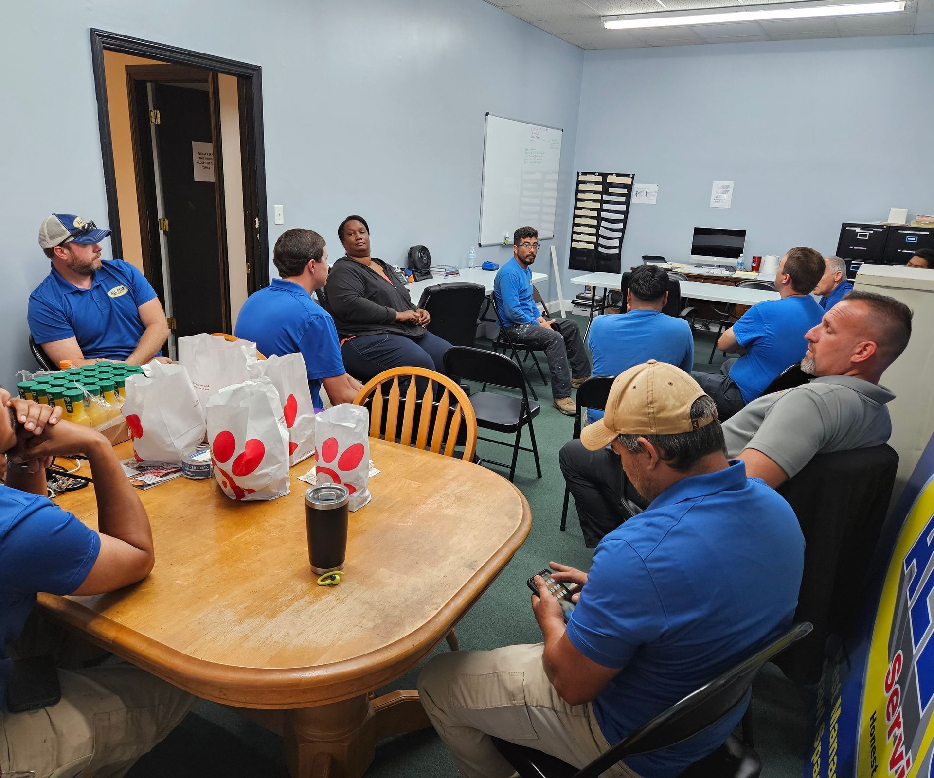 A group of people sit around a wooden table in an office, having a meal with Chick-fil-A takeout bags.