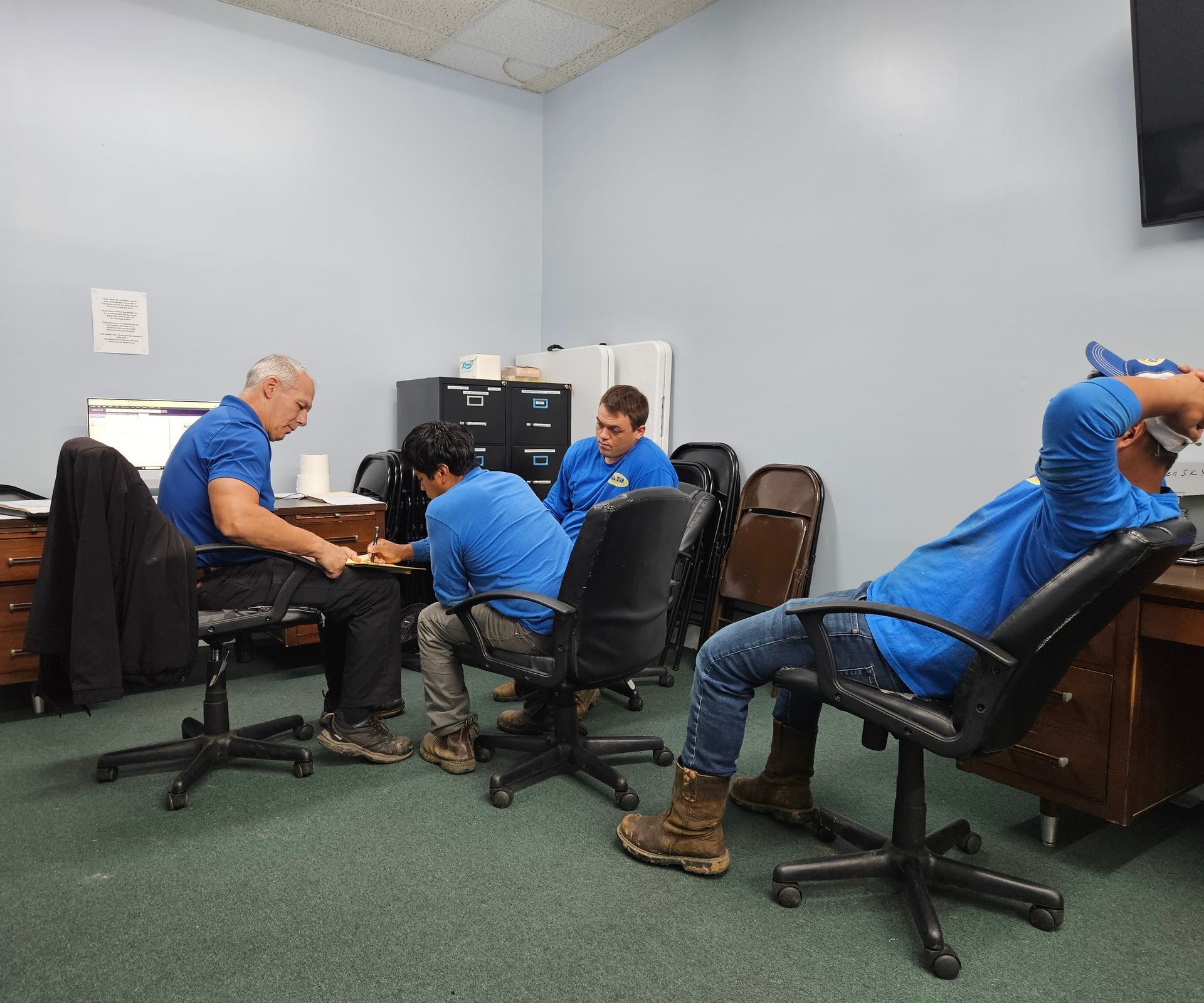 Four people in blue long-sleeve shirts sit in an office, with three focused on paperwork at a desk and one leaning back.
