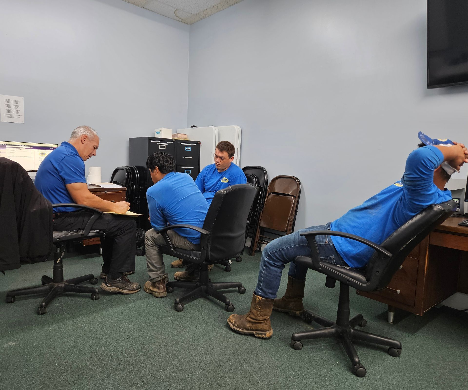 Four people in blue shirts are gathered for a meeting in an office, with some seated at desks and chairs.
