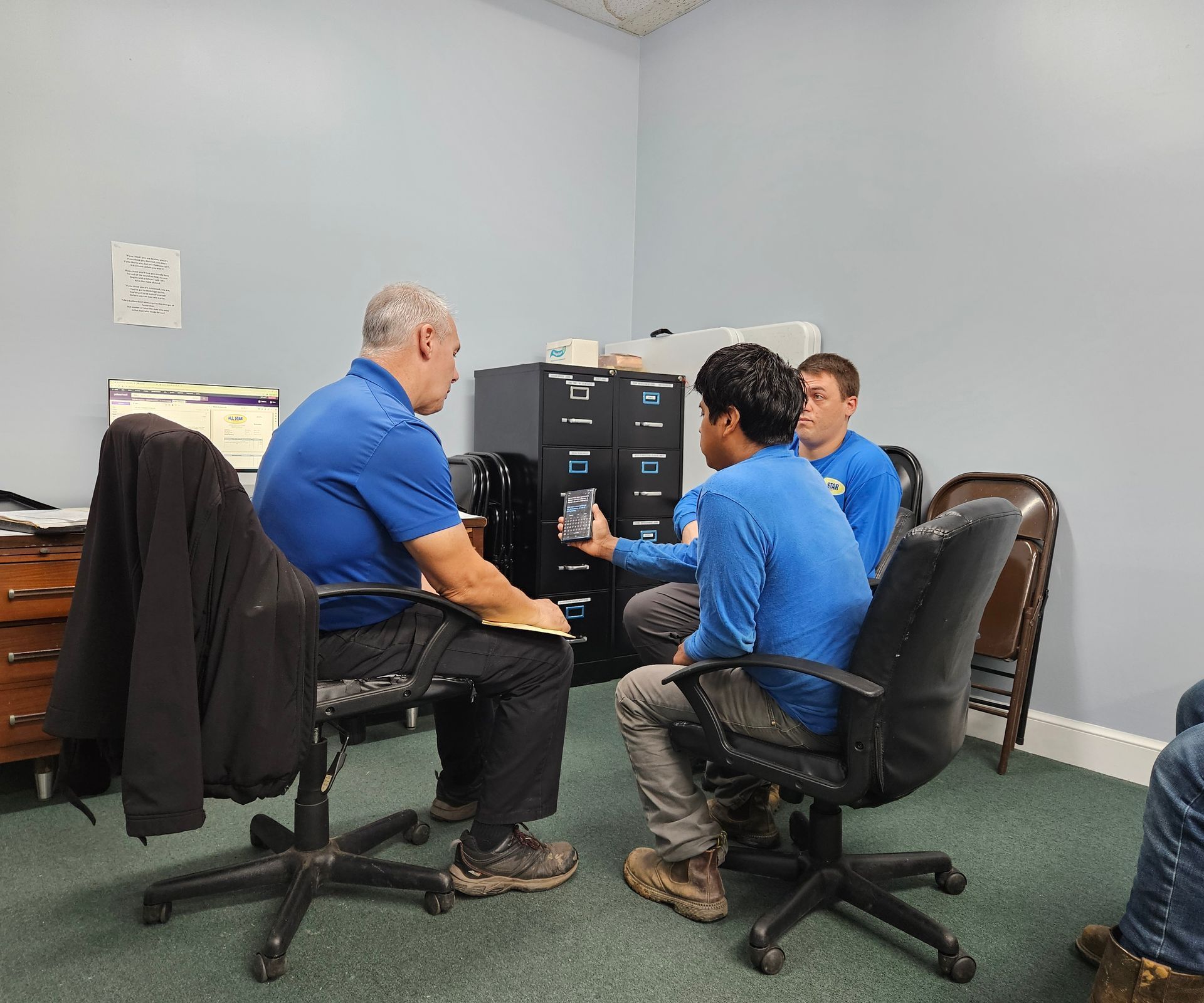 A man in a blue shirt discusses something with two seated individuals in an office setting with file cabinets.
