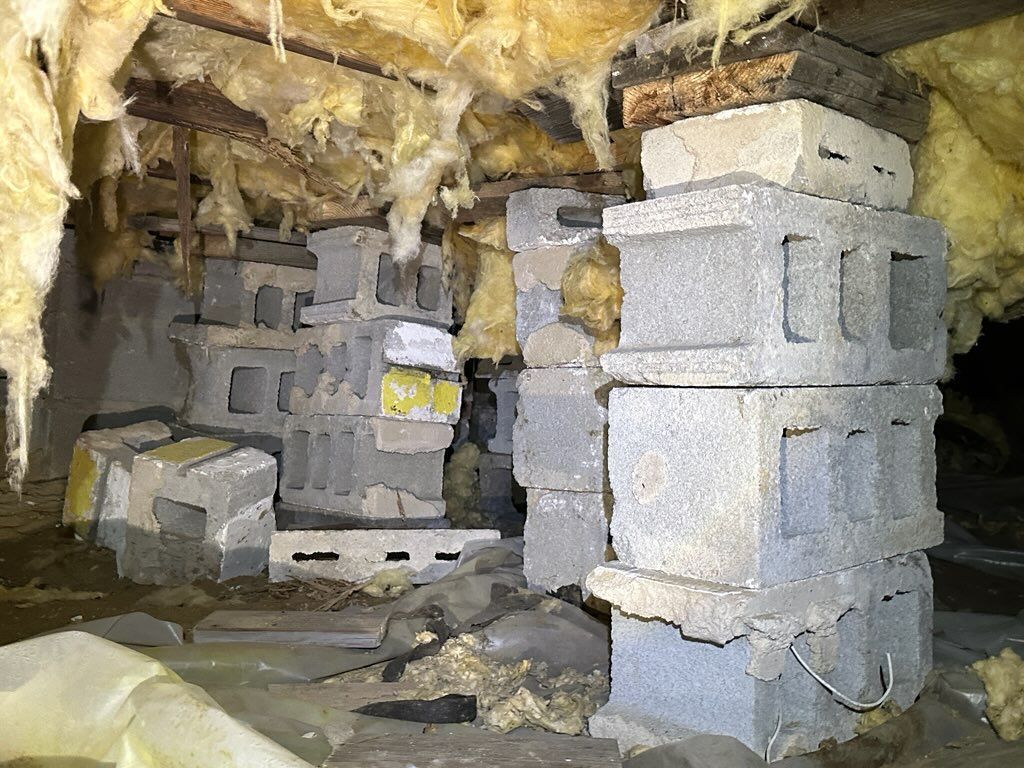 A crawlspace with sagging floor joists supported by unstable, dry-stacked concrete blocks.
