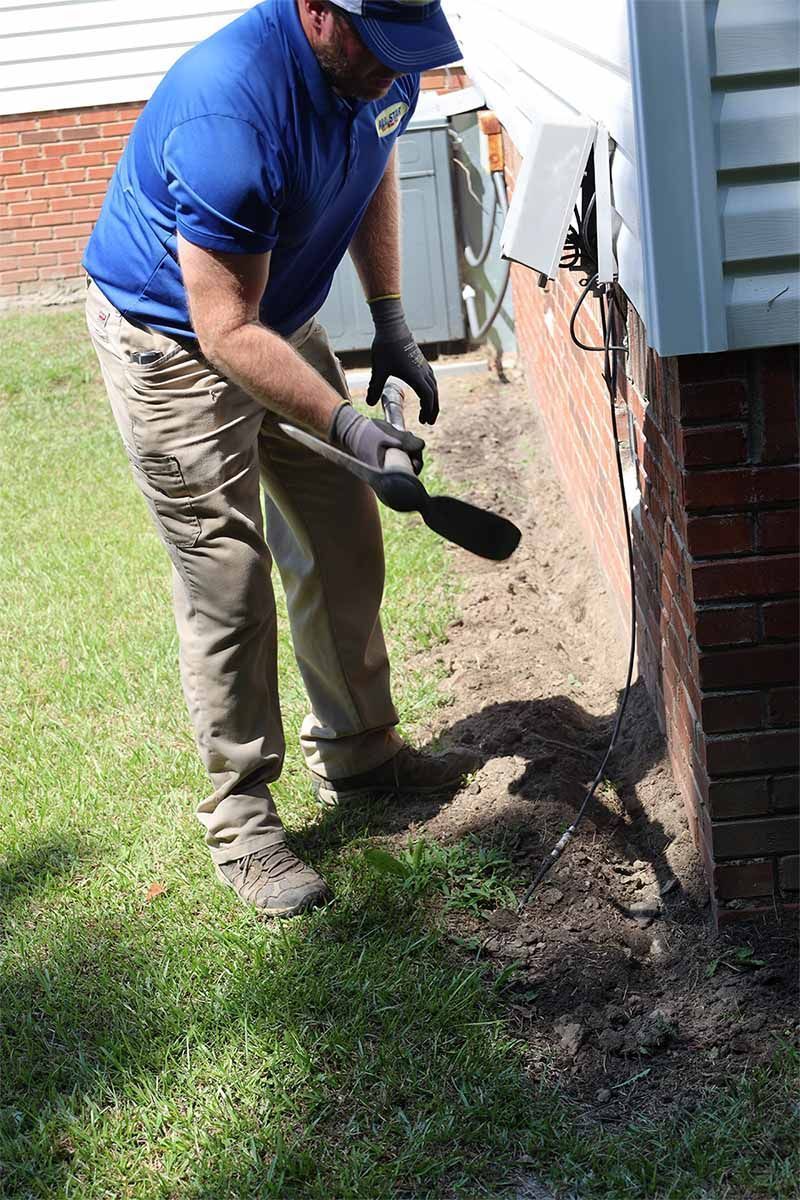 A person in a blue shirt and khaki pants uses a handheld digging tool along the edge of a brick house foundation.
