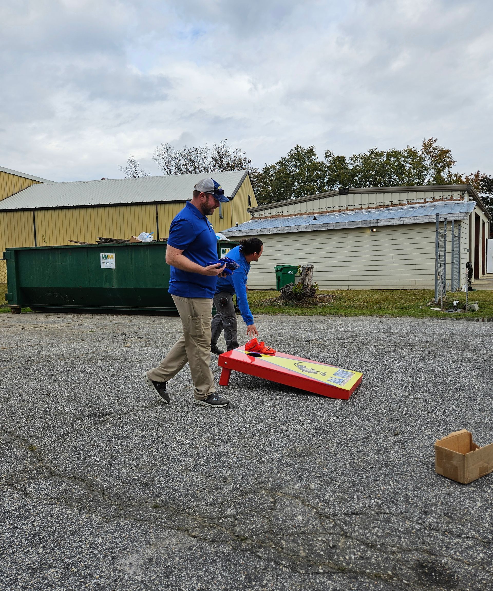 Two people set up a cornhole board on a gravel surface outdoors next to a green dumpster and old buildings.