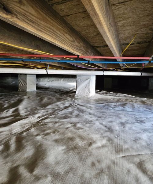 A low-angle view of a crawlspace with a white moisture barrier on the ground, support piers, and utility pipes overhead.