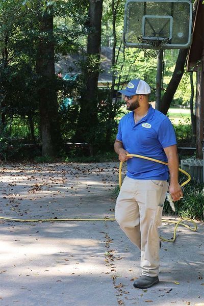 A person in a blue polo shirt and tan pants walks outdoors carrying a yellow garden hose on a paved driveway.