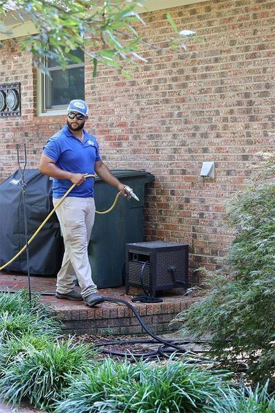 A technician wearing a blue polo and hat sprays a brick house exterior with a hose on a patio.