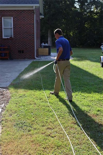 A person wearing a blue shirt and khaki pants uses a hose to spray water on a lawn near a brick house.