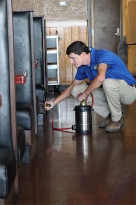 A person in a blue shirt and khakis crouches in a restaurant, using a stainless steel sprayer to treat booth seating.
