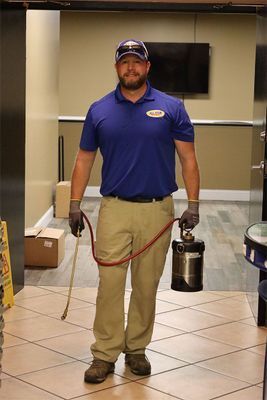 A pest control technician in a blue polo and khakis holds a metal pesticide sprayer in a building lobby.