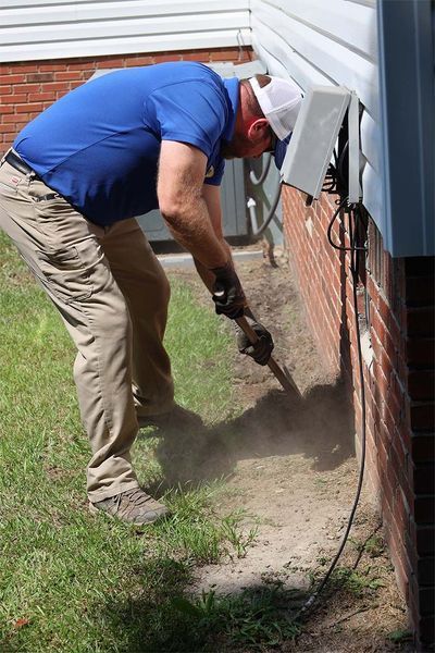 A person in a blue shirt and khaki pants digs in the dirt near a brick house foundation under an electrical box.