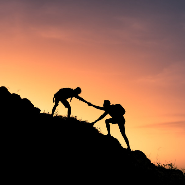 Two hikers silhouetted on a rocky slope at sunset, one helping the other climb