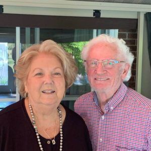 A woman with blonde hair and a pearl necklace standing next to a man with white hair and glasses, both smiling.