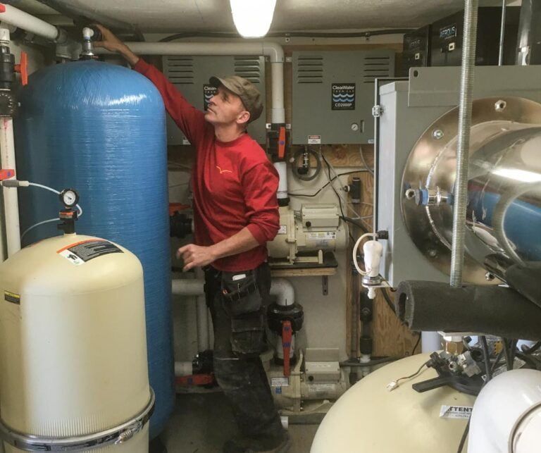 A technician in a red shirt works on plumbing and water filtration equipment in a mechanical room.