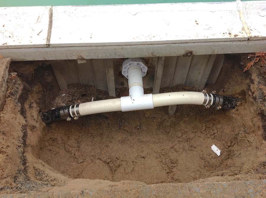 Un raccord en T en PVC blanc avec des tuyaux flexibles, partiellement enterré dans une tranchée de terre à côté du mur d'une piscine.