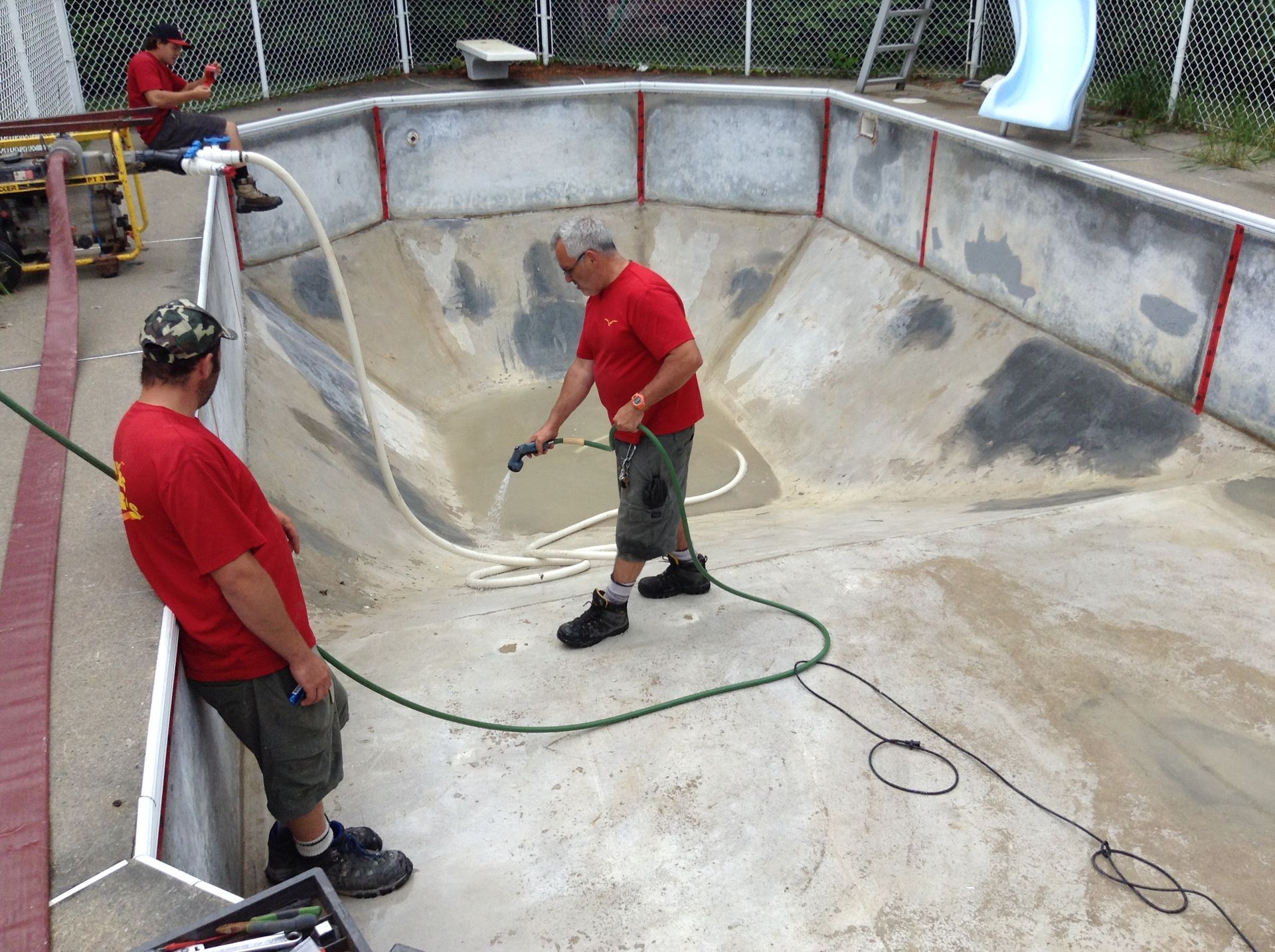 Deux ouvriers en chemises rouges nettoient une piscine en béton vide avec des tuyaux d'arrosage et des nettoyeurs haute pression.