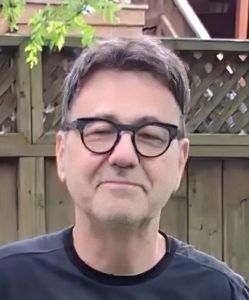 A person wearing dark-rimmed glasses and a black shirt, smiling slightly while standing in front of a wooden fence.