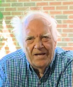 An older man with white hair wearing a blue and green plaid shirt, standing in front of a brick wall.
